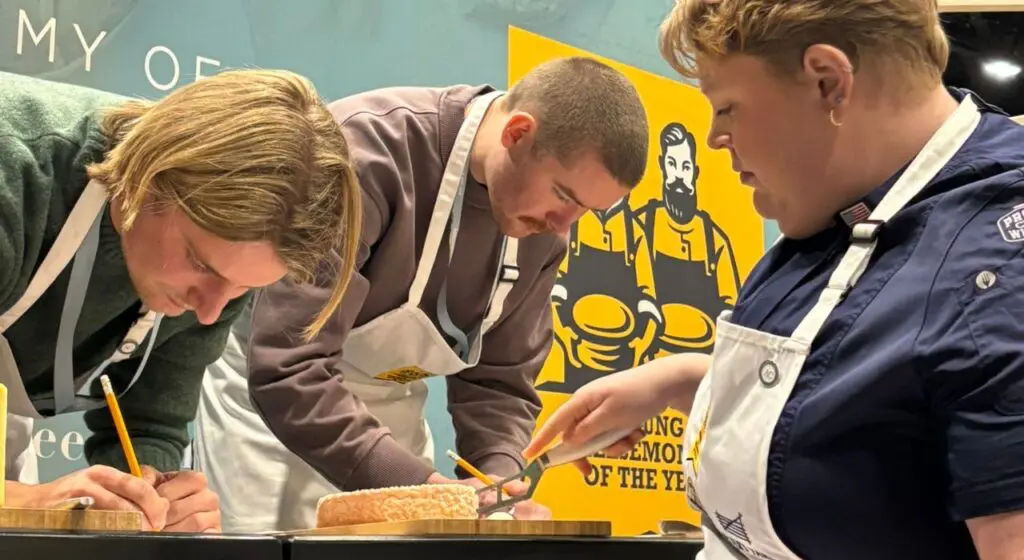 Three people wearing white aprons lean over a wooden cutting board, closely examining and working with cheese, with the Young Cheesemonger of the Year logo visible on a yellow poster in the background.