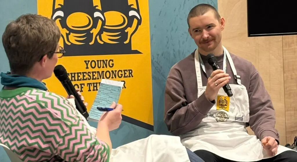 Two people sit in chairs having a conversation with microphones at an interview or panel event, with the Young Cheesemonger of the Year logo on a yellow poster visible behind them, one person wearing a green and white chevron-patterned sweater holding questions