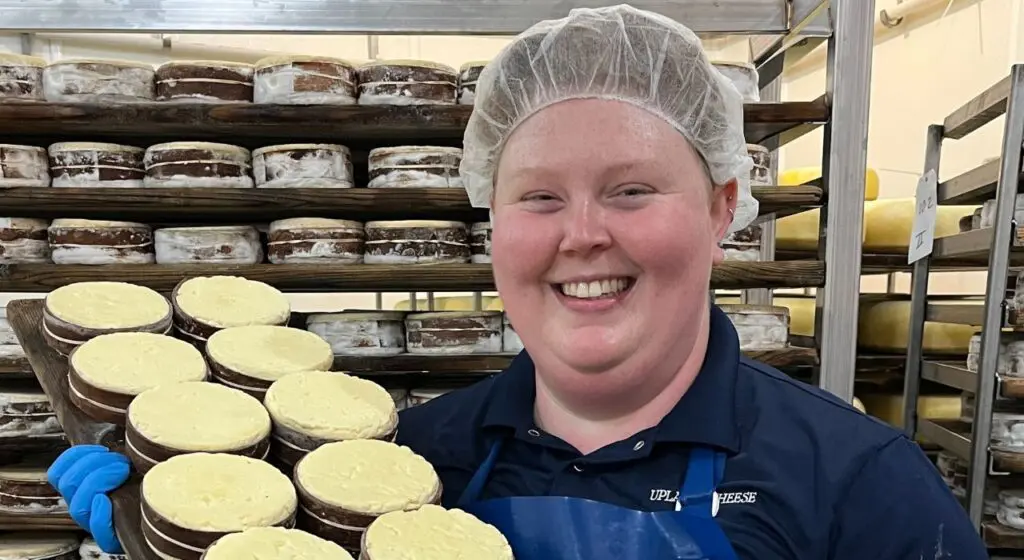 A person wearing a white hairnet and navy blue shirt smiles at the camera inside a cheese aging room, standing next to shelves filled with rounds of aging cheese wheels.