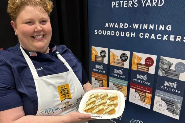 A smiling person wearing a white apron with a cheese competition logo holds a plate of crackers topped with cheese, standing in front of a display featuring Peter's Yard award-winning sourdough crackers packaging.