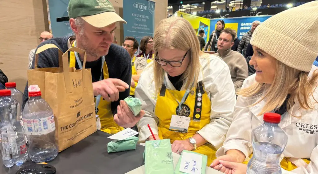 Three people wearing yellow aprons lean over a table at a busy trade show, examining and writing notes on small pieces of green paper, with water bottles and a shopping bag visible on the table.