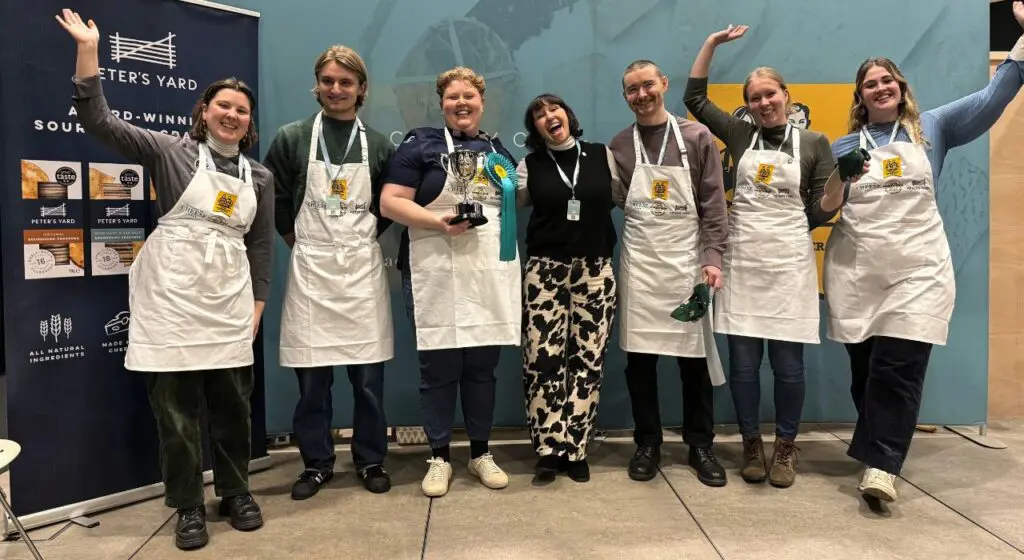 A group of seven people stand together smiling and cheering in front of event banners. Most are wearing white aprons with lanyards around their necks. One person in the center holds a trophy and wears award ribbons. The group appears celebratory and joyful, posing with raised arms.