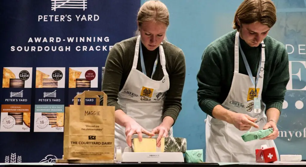 Two people wearing white aprons and green sweaters stand at a demonstration table in front of a Peter's Yard sourdough crackers display, cutting or wrapping cheese samples with a branded The Courtyard Dairy paper bag on the table.