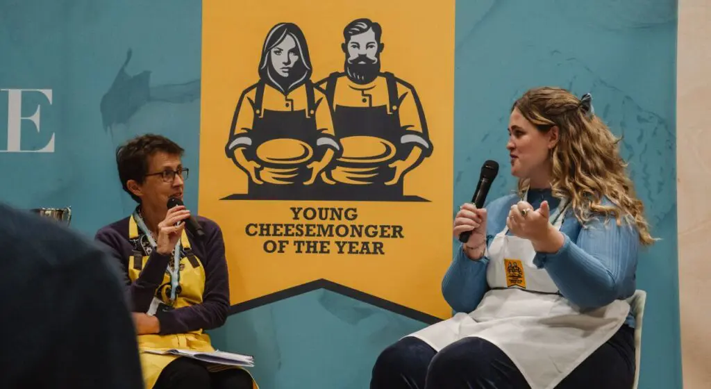 Two people sit in chairs having a conversation with microphones, with a large yellow poster behind them featuring an illustrated logo reading "Young Cheesemonger of the Year" showing two figures in aprons holding plates.