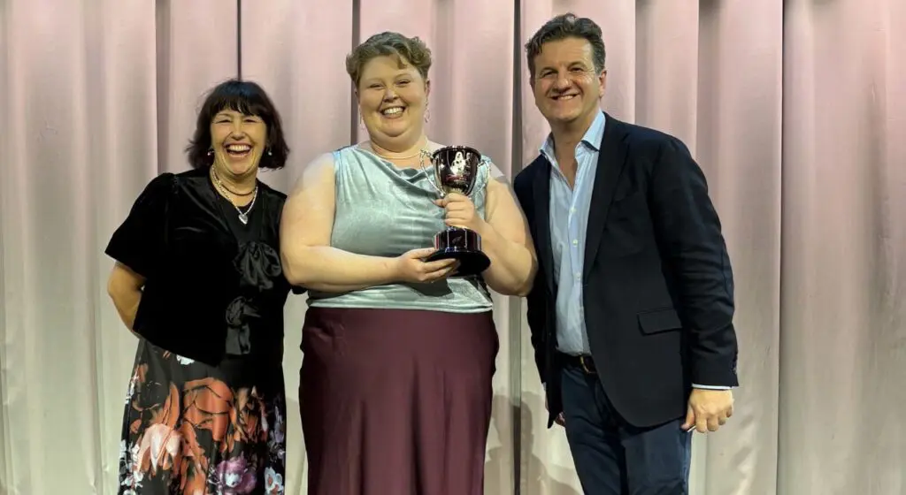 Three people pose together smiling in front of a white curtain backdrop, with the person in the center wearing a light blue top and burgundy skirt while holding a trophy with a cheesemonger figure on top.