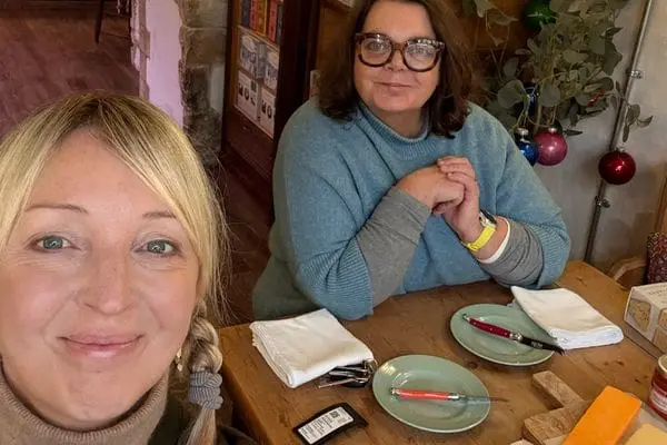 Two ladies sat at a wooden table looking at the camera, with two green plates and knives and a selection of cheeses on a board.