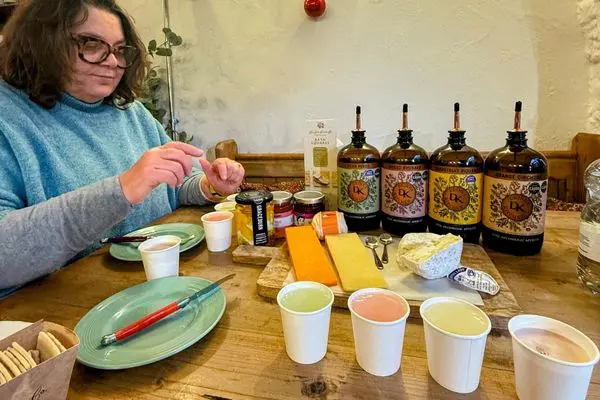 A woman in a blue jumper choosing from a selection of four non-alcoholic drinks in paper cups and a selection of cheese on a board. There is a green plate and knife in the image and four bottles in the background