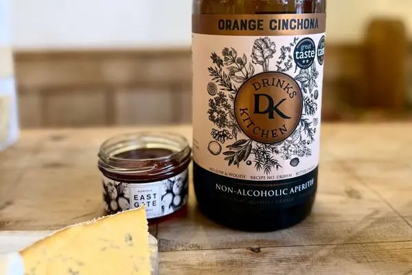 A bottle of orange cinchona non-alcoholic aperitif with a small jar of chutney and a slice of blue cheese.