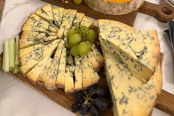A wedge of Blue Stilton on a wooden board with slices of stilton, green grapes, black grapes and celery