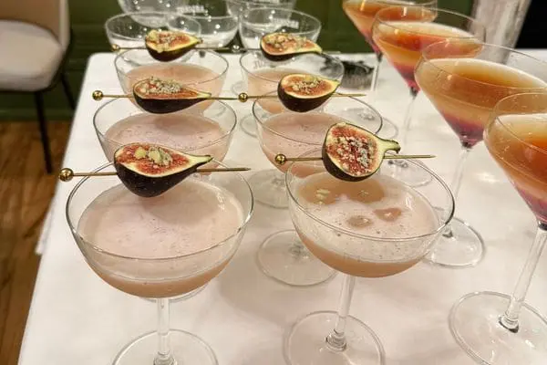 White clothed table with a number of coupe glasses with two different types of cocktail with figs on the top