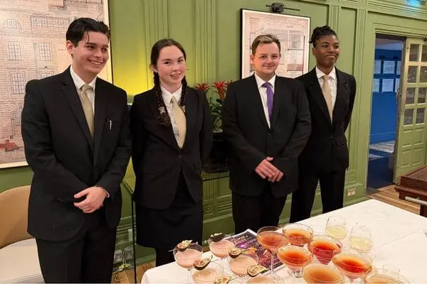 Four people in black suits and ties standing in front of a table with cocktails on.