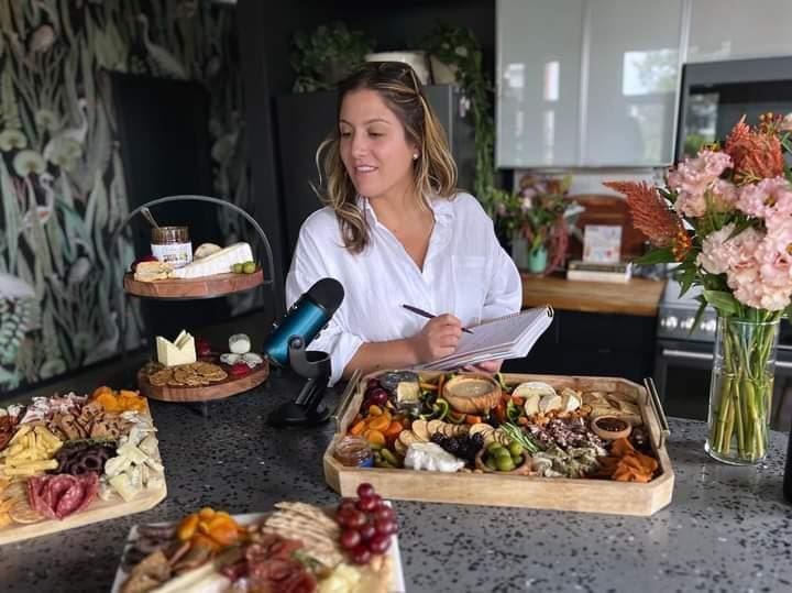 Brittany Bassett broadcasting her Curd Is The Word Podcast, in front of several boards filled with cheese and accompaniments